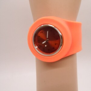 May include: Orange silicone wristwatch with a round, silver-rimmed face and black hour and minute hands.