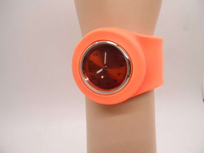 May include: Orange silicone wristwatch with a round, silver-rimmed face and black hour and minute hands.