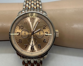 May include: A gold-toned watch with a silver band. The watch face has a rose gold background with black markings and three smaller dials.