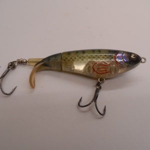 May include: A clear plastic fishing lure with a green and brown fish pattern. The lure has a tail that moves when it is in the water. It has two treble hooks.
