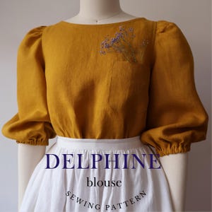 Könnte beinhalten: Eine senfgelbe Leinenbluse mit Puffärmeln und einer einzigen Tasche auf der linken Seite. Die Bluse hat einen runden Ausschnitt und wird mit einem weißen Rock kombiniert. Der Text "DELPHINE blouse SEWING PATTERN" ist auf dem Rock aufgedruckt.