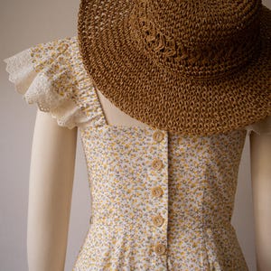 Puede incluir: Un vestido sin mangas de tela blanca con un peque&ntilde;o estampado floral amarillo, una hilera de botones de color marr&oacute;n claro en el centro y mangas con volantes y ribete de encaje blanco, junto a un sombrero de paja trenzado marr&oacute;n.