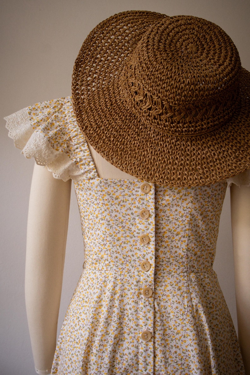 Puede incluir: Un vestido sin mangas de tela blanca con un peque&ntilde;o estampado floral amarillo, una hilera de botones de color marr&oacute;n claro en el centro y mangas con volantes y ribete de encaje blanco, junto a un sombrero de paja trenzado marr&oacute;n.
