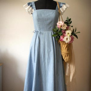 Puede incluir: Un vestido midi azul claro con escote cuadrado y mangas con volantes y ribete de encaje blanco. El vestido presenta un corpi&ntilde;o ajustado con finas cintas laterales en la cintura y una falda acampanada. Un bolso tejido beige con flores cuelga de un lado.