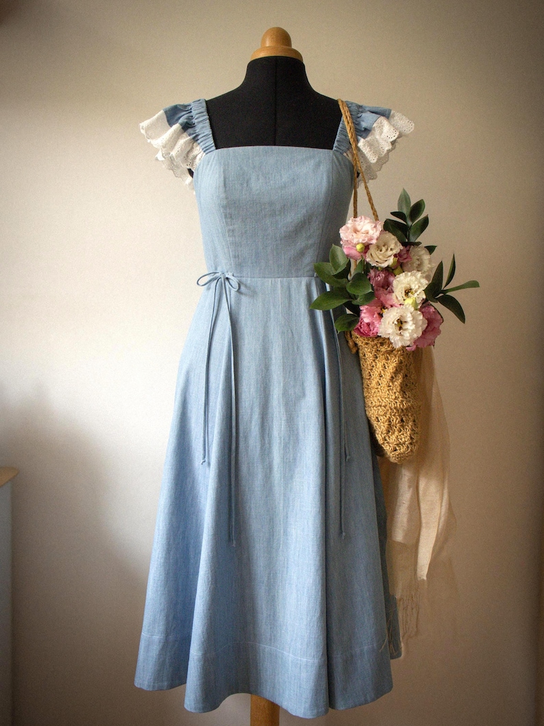 Puede incluir: Un vestido midi azul claro con escote cuadrado y mangas con volantes y ribete de encaje blanco. El vestido presenta un corpi&ntilde;o ajustado con finas cintas laterales en la cintura y una falda acampanada. Un bolso tejido beige con flores cuelga de un lado.