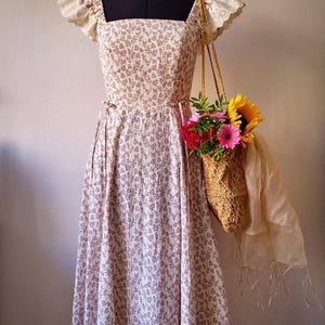 Puede incluir: Un vestido midi con escote cuadrado y mangas cortas de encaje de bolillo color crema. El vestido presenta un estampado floral marr&oacute;n sobre un fondo claro. Un bolso de paja trenzada lleno de girasoles y flores rosas cuelga del hombro.