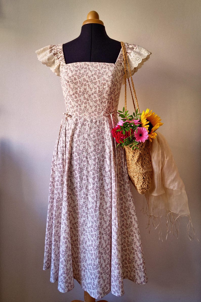 Puede incluir: Un vestido midi con escote cuadrado y mangas cortas de encaje de bolillo color crema. El vestido presenta un estampado floral marr&oacute;n sobre un fondo claro. Un bolso de paja trenzada lleno de girasoles y flores rosas cuelga del hombro.