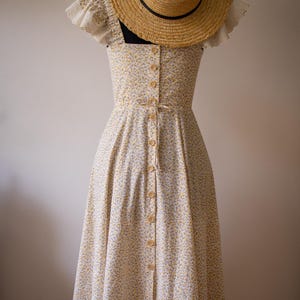 Puede incluir: Un vestido midi con un peque&ntilde;o estampado floral amarillo y azul sobre fondo blanco, con escote cuadrado, mangas cortas con volantes, una hilera de botones frontal y un lazo en la cintura, acompa&ntilde;ado de un sombrero de paja de ala ancha.