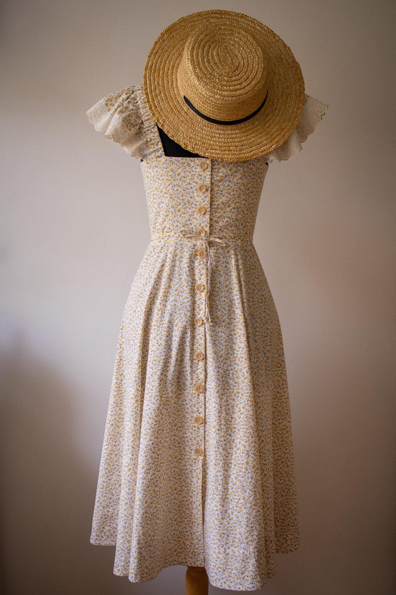 Puede incluir: Un vestido midi con un peque&ntilde;o estampado floral amarillo y azul sobre fondo blanco, con escote cuadrado, mangas cortas con volantes, una hilera de botones frontal y un lazo en la cintura, acompa&ntilde;ado de un sombrero de paja de ala ancha.