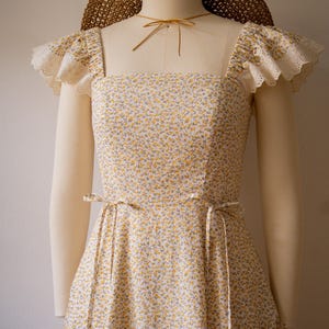 Puede incluir: Un vestido sin mangas con un peque&ntilde;o estampado floral amarillo y verde sobre fondo blanco, escote cuadrado y mangas con volantes de encaje de ojal color crema. El vestido incluye finas cintas en la cintura y se combina con un sombrero de paja trenzada marr&oacute;n.