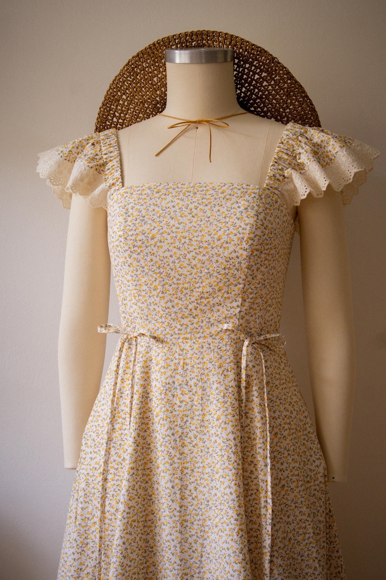 Puede incluir: Un vestido sin mangas con un peque&ntilde;o estampado floral amarillo y verde sobre fondo blanco, escote cuadrado y mangas con volantes de encaje de ojal color crema. El vestido incluye finas cintas en la cintura y se combina con un sombrero de paja trenzada marr&oacute;n.