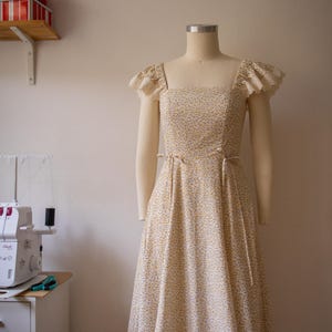 Puede incluir: Un vestido midi con un peque&ntilde;o estampado floral amarillo y azul sobre un fondo crema. El vestido tiene un escote cuadrado, mangas con volantes y ribete de encaje, y finas cintas de tela en la cintura, expuesto sobre un maniqu&iacute; de costura.