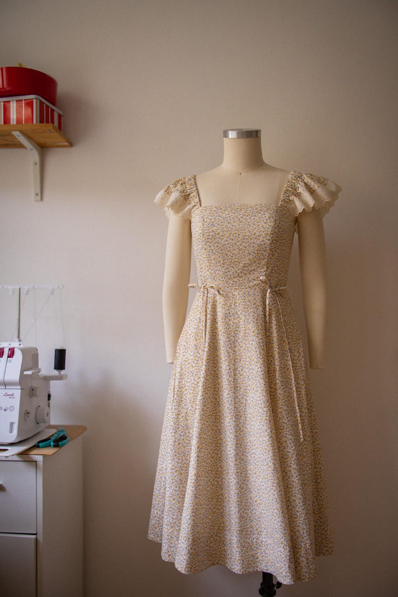 Puede incluir: Un vestido midi con un peque&ntilde;o estampado floral amarillo y azul sobre un fondo crema. El vestido tiene un escote cuadrado, mangas con volantes y ribete de encaje, y finas cintas de tela en la cintura, expuesto sobre un maniqu&iacute; de costura.
