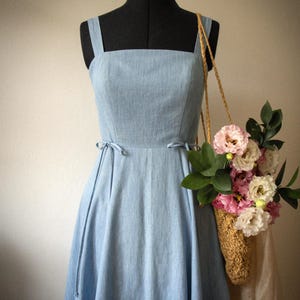 Puede incluir: Un vestido azul claro sin mangas con escote cuadrado y finos lazos laterales en la cintura. El vestido se muestra en un maniqu&iacute; junto a un bolso de paja trenzada lleno de flores rosas y blancas y hojas verdes, colgado del hombro.