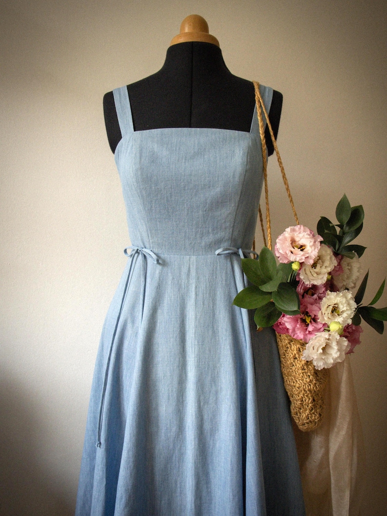 Puede incluir: Un vestido azul claro sin mangas con escote cuadrado y finos lazos laterales en la cintura. El vestido se muestra en un maniqu&iacute; junto a un bolso de paja trenzada lleno de flores rosas y blancas y hojas verdes, colgado del hombro.