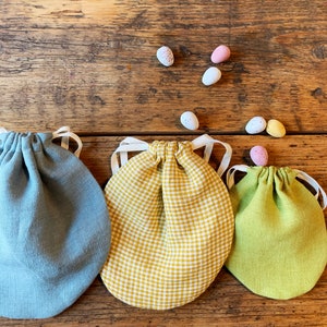 May include: Three small fabric drawstring bags in different colors and patterns. The bags are on a wooden surface with small candy eggs scattered around them.