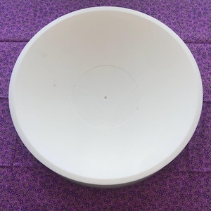 May include: A white, round bowl with a wide, shallow design. The bowl has a smooth interior and a slightly textured exterior. The centre of the bowl features a small, circular indentation. The bowl is set against a purple patterned background.