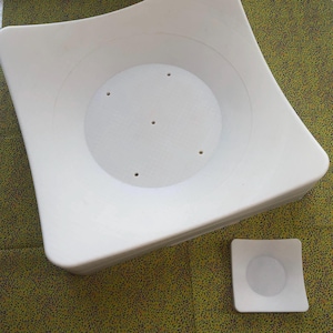 Op de afbeelding: Twee witte vierkante taartvormen van plastic met een verhoogd midden en afvoergaten. De kleinere vorm ligt op de grotere vorm.