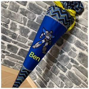 Könnte beinhalten: Ein blauer und schwarzer Schultüte mit einem Roboter-Design und dem Namen "Ben" bestickt. Die Tüte ist mit einem gelben Band verziert.