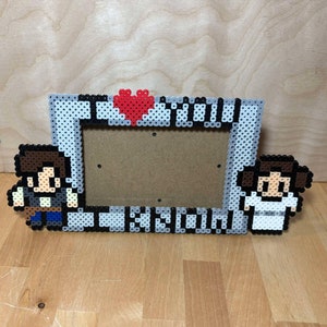 May include: A gray and white pixel art picture frame with a red heart in the top center. The frame features a pixel art image of Han Solo and Princess Leia on either side of the frame.