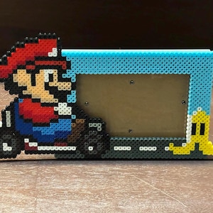 May include: A handmade photo frame featuring a pixelated design of Mario in a kart, crafted from colorful plastic beads. The frame has a light blue border and a yellow banana peel accent. The frame is approximately 8 inches wide.