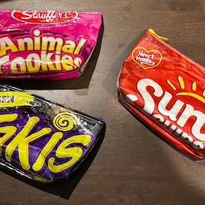 May include: Three zippered pouches made from repurposed snack packaging. One pink pouch features "Animal Cookies" text, a red pouch displays "Sun" and a yellow sun graphic, and a purple pouch has "Takis" in yellow.