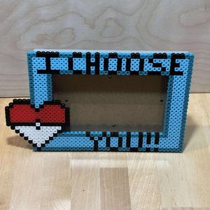 May include: A blue and black beaded picture frame with a red and white heart design. The frame says "I Choose You" in black beads.