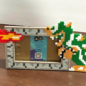 May include: A pixel art photo frame featuring a gray brick-like border. The frame is decorated with a red and yellow flame on the left and a green and orange character on the right. The frame has a brown backing and a blue label with text.