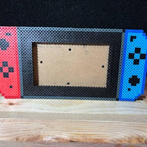 May include: A pixel art picture frame shaped like a Nintendo Switch console. The frame is made of black, red, and blue beads. The frame is empty and ready for a photo.