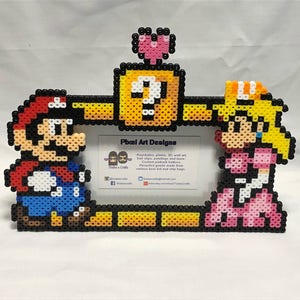 Könnte beinhalten: Ein Pixel-Art-Bilderrahmen mit Mario und Prinzessin Peach aus der Super Mario Bros.-Videospielserie. Der Rahmen besteht aus bunten Plastikperlen und enthält oben eine herzförmige Perle. Der Rahmen ist mit einem gelben Fragezeichenblock und einer gelben Plattform verziert. Der Rahmen ist von einem schwarzen Rand umgeben. Der Rahmen enthält eine Visitenkarte mit dem Text "Pixel Art Designs".