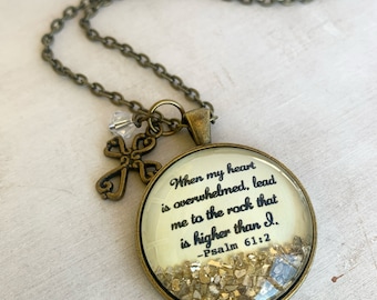 May include: A gold necklace with a round pendant featuring the text "When my heart is overwhelmed, lead me to the rock that is higher than I. -Psalm 61:2" and gold glitter inside. The necklace also has a small cross charm.
