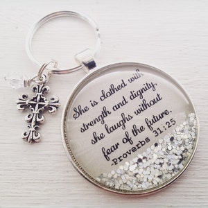 May include: A silver keychain with a round glass charm that says "She is clothed with strength and dignity, she laughs without fear of the future. -Proverbs 31:25" and silver glitter inside. The charm is attached to a silver key ring with a small silver cross charm.