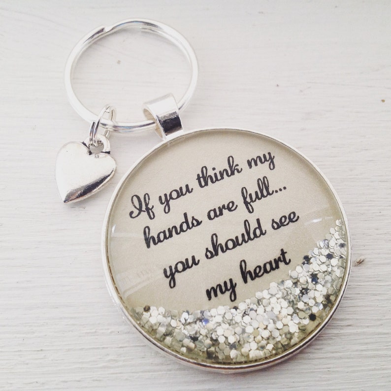 May include: Silver keychain with a heart charm and a round glass charm with silver glitter. The glass charm has the text "If you think my hands are full... you should see my heart".
