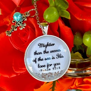 May include: A silver necklace with a round pendant featuring a white background with black text that reads "Mightier than the waves of the sea is His love for you - Psalm 93:4". The pendant is surrounded by silver glitter. The necklace also features a silver cross charm with a light blue bead.