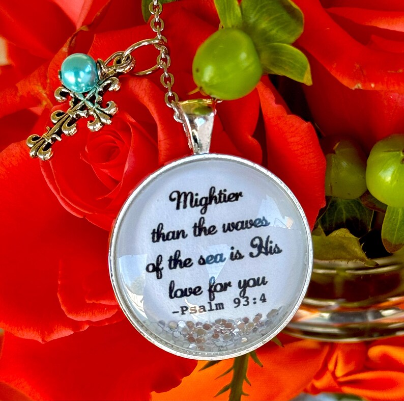 May include: A silver necklace with a round pendant featuring a white background with black text that reads "Mightier than the waves of the sea is His love for you - Psalm 93:4". The pendant is surrounded by silver glitter. The necklace also features a silver cross charm with a light blue bead.