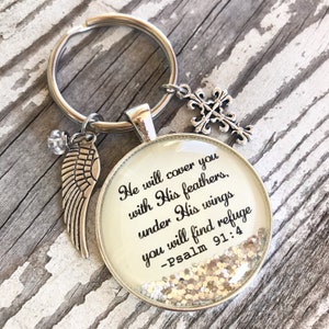 以下が含まれることがあります： 「He will cover you with His feathers, under His wings you will find refuge -Psalm 91:4」と書かれた丸いガラス製のチャームが付いたシルバーのキーホルダー。チャームの中には金色のグリッターが入っています。キーホルダーには、シルバーの天使の羽根のチャームとシルバーの十字架のチャームも付いています。