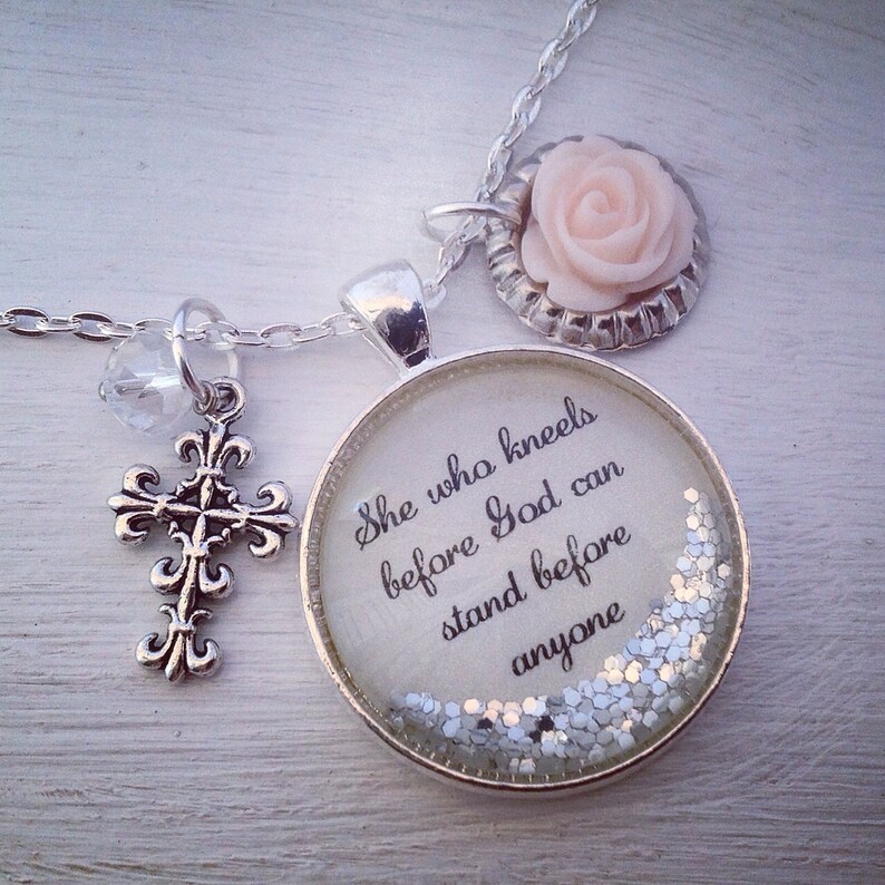 Puede incluir: Collar de plata con un dije de cruz, un peque&ntilde;o dije de cristal y un dije redondo con la cita "Ella que se arrodilla ante Dios puede estar de pie ante cualquiera" y purpurina plateada en su interior.