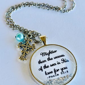 May include: A silver necklace with a round pendant featuring the quote "Mightier than the waves of the sea is His love for you - Psalm 93:4" and a small silver cross charm. The pendant has a clear background with silver glitter.