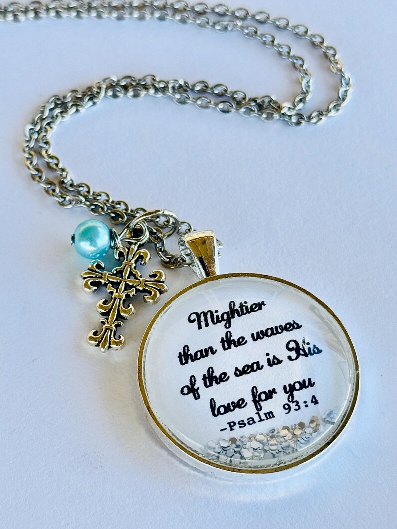May include: A silver necklace with a round pendant featuring the quote "Mightier than the waves of the sea is His love for you - Psalm 93:4" and a small silver cross charm. The pendant has a clear background with silver glitter.