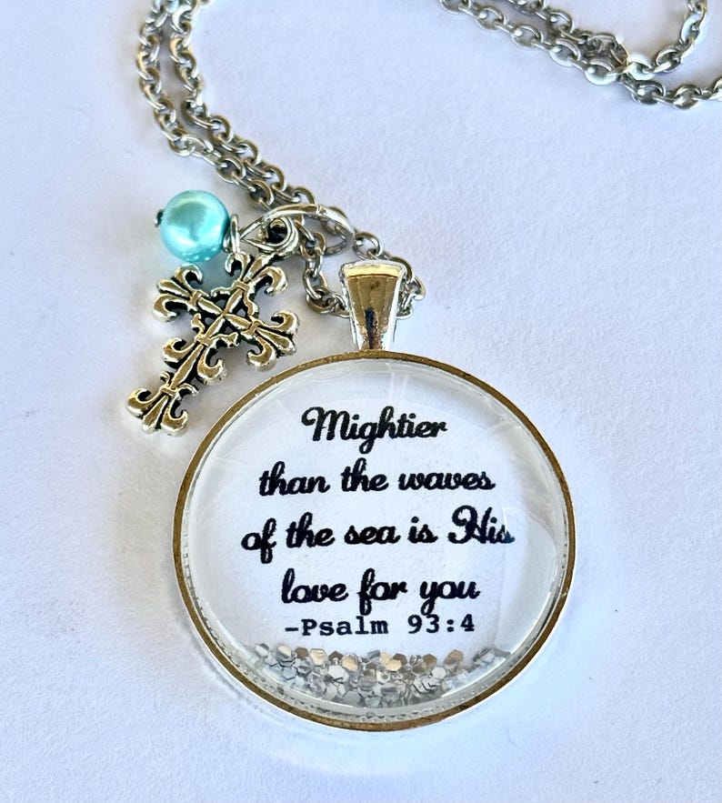 May include: A silver necklace with a round glass pendant that says "Mightier than the waves of the sea is His love for you - Psalm 93:4". The pendant has silver glitter inside. The necklace also has a silver cross charm and a light blue bead.