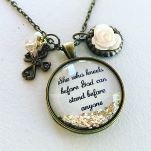 Puede incluir: Un collar de oro con un colgante de vidrio que dice "Ella que se arrodilla ante Dios puede estar de pie ante cualquiera". El colgante tiene purpurina dorada en su interior. El collar tambi&eacute;n tiene un peque&ntilde;o dije de cruz y un dije de rosa blanca.