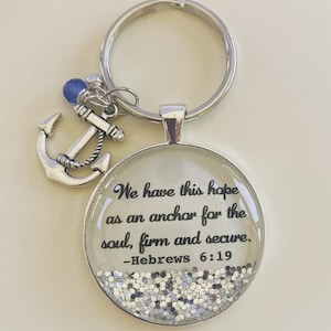 May include: Silver keychain with a small anchor charm and a round glass charm with glitter. The glass charm has the text "We have this hope as an anchor for the soul, firm and secure. -Hebrews 6:19"