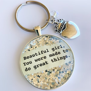 May include: Silver keychain with a round pendant containing the text "Beautiful girl, You were made to do great things." The pendant is surrounded by silver glitter. A crown and heart charm dangle from the keychain.