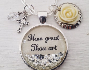 May include: Silver necklace with a round pendant that says "How great Thou art" and has silver glitter inside. The necklace also has a small cross charm and a white flower charm.