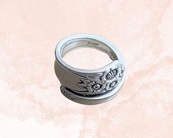 Op de afbeelding: Een zilverkleurige ring gemaakt van een lepel, met bloemendetails op de voorkant. De ring heeft een gebogen ontwerp, waarbij de steel van de lepel de band vormt. De ring is geplaatst tegen een zachte, roze en witte achtergrond.