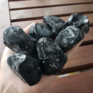 Pode incluir: Um punhado de pedras de obsidiana preta polidas. As pedras têm uma superfície lisa e brilhante com algumas veias brancas. As peças de obsidiana variam em tamanho e forma, com bordas arredondadas.
