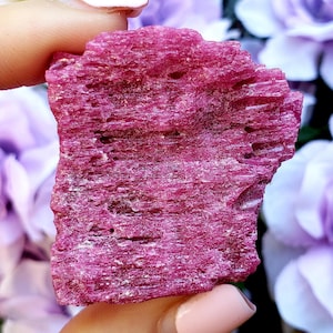 May include: A raw ruby gemstone in a deep pink shade. The uncut mineral displays a textured surface with visible crystalline structures and natural flaws. Soft, blurred purple flowers form the background.