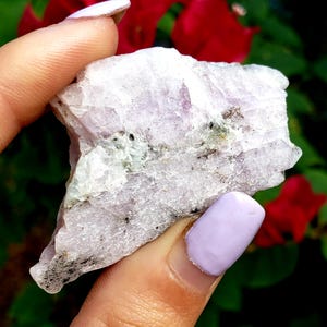May include: A raw, light purple and white crystal with a rough, natural texture. The crystal has a slightly translucent appearance and is speckled with small, dark gray inclusions.