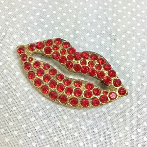 Pode incluir: Um broche dourado em forma de lábios, incrustado com strass vermelhos.