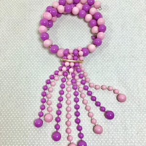 May include: A purple and pink beaded necklace with a tassel. The necklace is made of round beads and has a gold clasp.